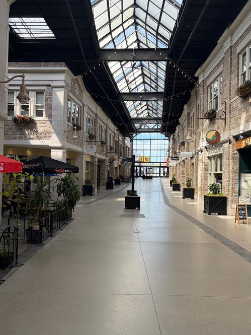Long indoor shopping arcade with glass skylights, string lights, and storefronts on both sides; potted plants line the walkway.