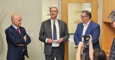 Three men in suits stand in an office hallway; center speaker holds papers while two colleagues listen, as a photographer on the right captures the moment.