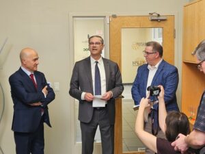 Three men in suits stand in an office as the central speaker holds papers; a photographer records the moment.
