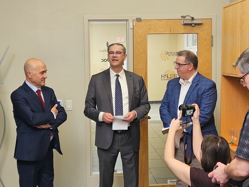 Three men in suits stand in an office as the central speaker holds papers; a photographer records the moment.