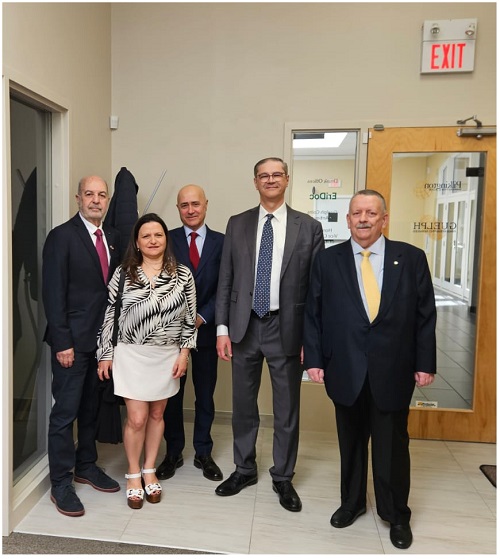 Five professionally dressed people pose for a photo in a hallway outside a glass-door entry, with an EXIT sign visible above.