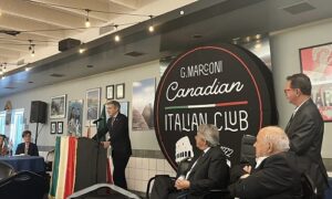 Man at a podium speaking during a Canadian Italian Club event, with an Italian flag draped on the front and seated attendees nearby.