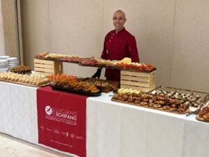 Chef in a red uniform stands behind a long buffet table with assorted pastries and appetizers on wooden crates at an event, bannered with Luciano Schipano Hospitality Group logo.