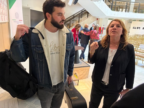 Bearded man with a denim jacket and backpack listens as a woman in a black blazer speaks in a bright, busy atrium; a guitar case rests nearby.
