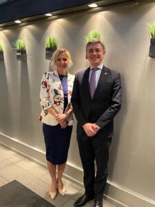 Two professionally dressed people stand side by side in a hallway with wall-mounted planters, smiling at the camera; the woman wears a floral blazer and navy dress, the man a dark suit and purple tie.