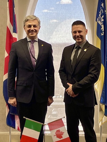 Two men in dark suits stand side by side indoors, with Italian and Canadian flags visible in front.