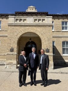 Three men in suits stand for a photo in front of a brick building with an arched entrance.