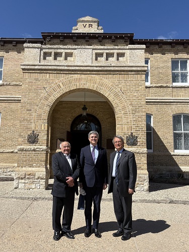 Three men in suits stand for a photo in front of a brick building with an arched entrance.