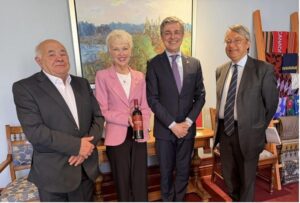 Four adults in business attire posing indoors for a group photo; the woman in a pink blazer holds a bottle as if presenting it, with three men standing beside her.