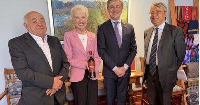 Four adults in business attire posing indoors for a group photo; the woman in a pink blazer holds a bottle as if presenting it, with three men standing beside her.