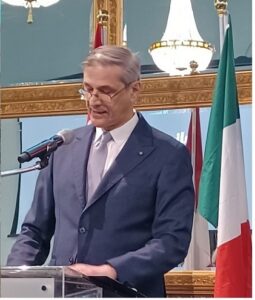 Middle-aged man in a dark suit speaks at a podium with a microphone, Italian flag in the background and ornate decor behind him.