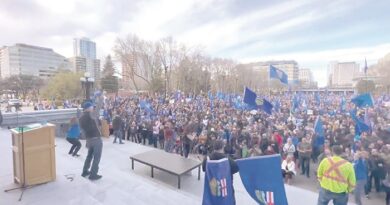 Large crowd at a rally with blue flags, gathered on steps outside a city hall or park, a speaker addressing them from a podium.