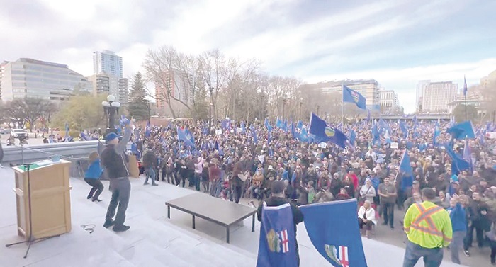 Large crowd at a rally with blue flags, gathered on steps outside a city hall or park, a speaker addressing them from a podium.