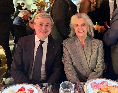 Smiling man in a dark suit and tie and an elderly woman in a silver blazer pose at a formal dinner table.