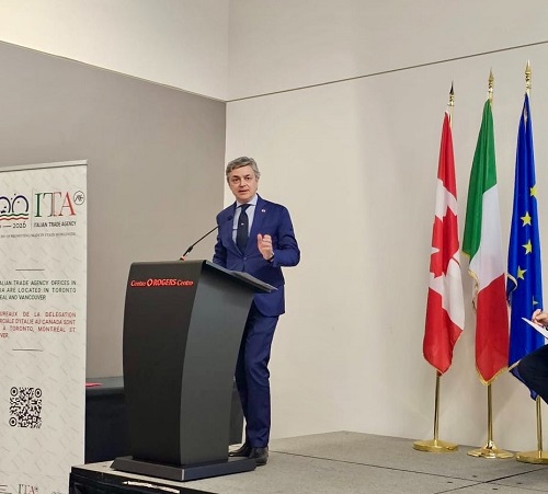 Man in a blue suit speaks at a conference podium on a stage with Swiss, Italian, and EU flags behind him.