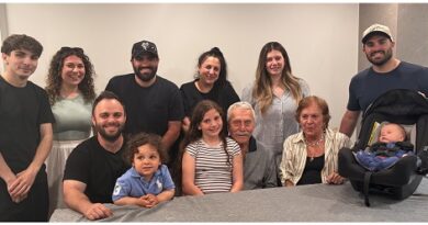 Large group of people of varying ages posing for a casual group photo around a table; two children sit at the front and a baby in a car seat is on the right.