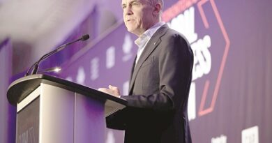 Man in a dark suit delivering a speech at a podium on a conference stage with purple lighting in the background.