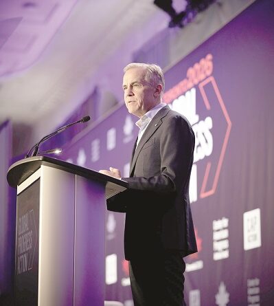Man in a dark suit delivering a speech at a podium on a conference stage with purple lighting in the background.