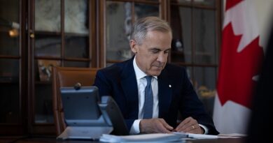 Man in a navy suit signing papers at a wooden desk in a formal office, with a Canadian flag behind him.