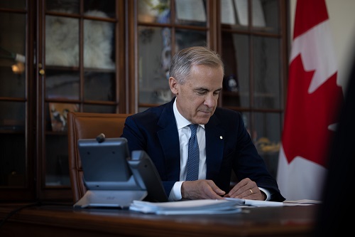 Man in a navy suit signing papers at a wooden desk in a formal office, with a Canadian flag behind him.