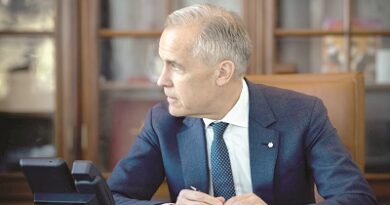 Man in a navy suit sits at a large wooden desk in an office, holding a pen and looking to the left, with a phone and papers on the desk.