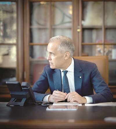 Man in a navy suit sits at a large wooden desk in an office, holding a pen and looking to the left, with a phone and papers on the desk.