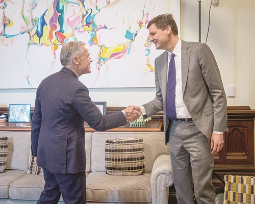 Two businesspeople in suits shake hands in a formal office with a colorful abstract painting in the background.