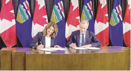 Two officials signing documents at a wooden table with Canadian and Alberta flags in the background.