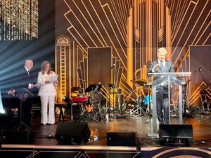 Man in a suit speaks at a clear lectern on a stage with a gold Art Deco backdrop and band setup behind him.