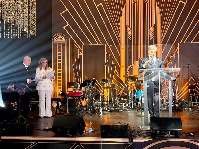 Man in a suit speaks at a clear lectern on a stage with a gold Art Deco backdrop and band setup behind him.