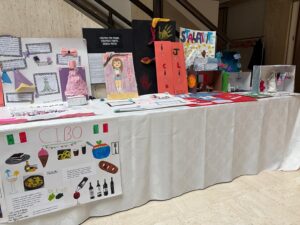 Table covered with a white cloth displaying student projects and dioramas, including colorful posters, handmade models, and a collage board with drawings and cutouts.
