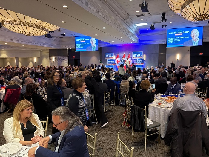 Crowded banquet hall at a conference with round tables; stage with speakers and screens showing Steven Del Duca.