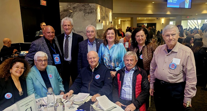 Group of eleven adults posing for a photo around a round banquet table at a conference, with name badges and formal attire in a busy event hall.