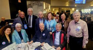Group of mostly older adults posing for a photo around a round banquet table at a conference event, some wearing name badges and lanyards.