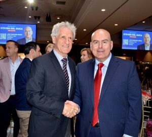 Two men in suits shake hands at a formal event, with guests and a Steven Del Duca campaign poster in the background.