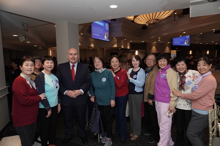 A diverse group of adults posing together with a man in a dark suit and red tie at an indoor event, all wearing circular badges on their clothes.
