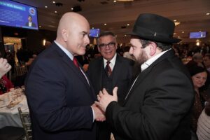 Two men in formal suits shake hands at a banquet; left is a bald man in a navy suit with a red tie, right is a bearded man in a black suit and hat, with a smiling man in glasses behind them.