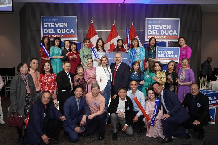 Group of supporters posing for a campaign photo indoors, with 'Re-Elect Steven Del Duca for Mayor' signs and Canadian flags behind them.