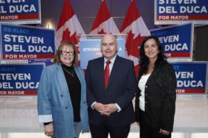 Three people pose at a Steven Del Duca for Mayor campaign event, with blue signs and Canadian flags behind them.