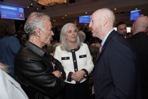 Three adults in a formal networking event chat closely: a man in a black leather jacket, a woman in a white blazer, and a man in a dark suit.