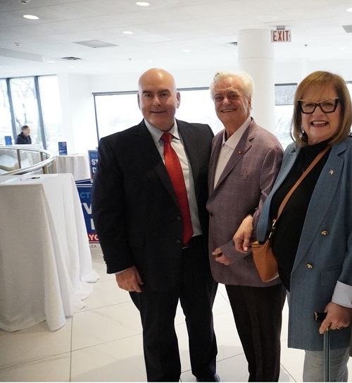 Three adults pose for a photo in a bright lobby: a man in a black suit and red tie, an older man in a purple blazer, and a woman wearing glasses and a blue coat.