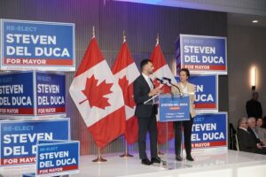 Man and woman stand at a podium on a campaign stage beside Canadian flags and multiple blue campaign signs reading 'RE-ELECT ... FOR MAYOR'.