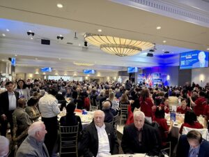 Large conference hall filled with attendees at round tables for a banquet-style event, stage and screens visible in the background.