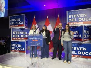 Group of campaign supporters on a stage with blue 'Steven Del Duca for Mayor' signs and Canadian flags in the background.
