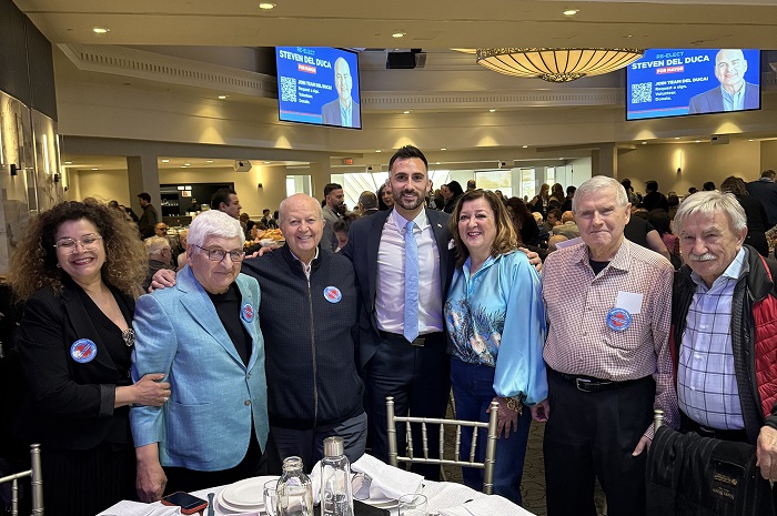 Eight people wearing name badges pose for a group photo at a banquet in a conference hall, with screens in the background displaying Steven Del Duca.