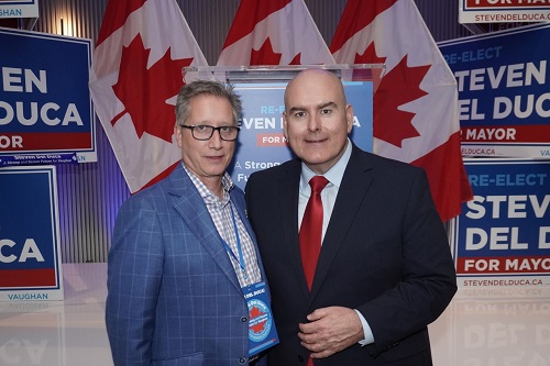 Two men in suits posing at a campaign event with Canadian flags and Steven Del Duca signs in the background.