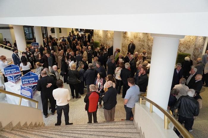 Crowd of people mingling in a spacious lobby by a stairway; campaign signs for Steven Del Duca visible at left and a table with signs nearby.