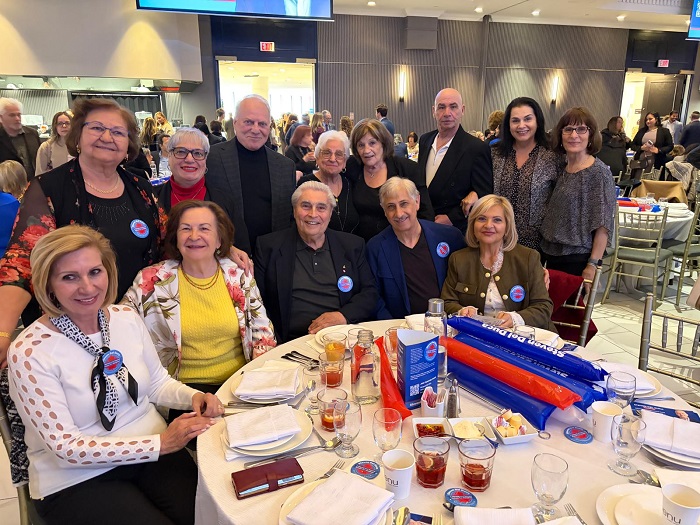 Twelve smiling adults posing around a round banquet table in a formal event hall, some wearing campaign stickers and drinks on the table behind them