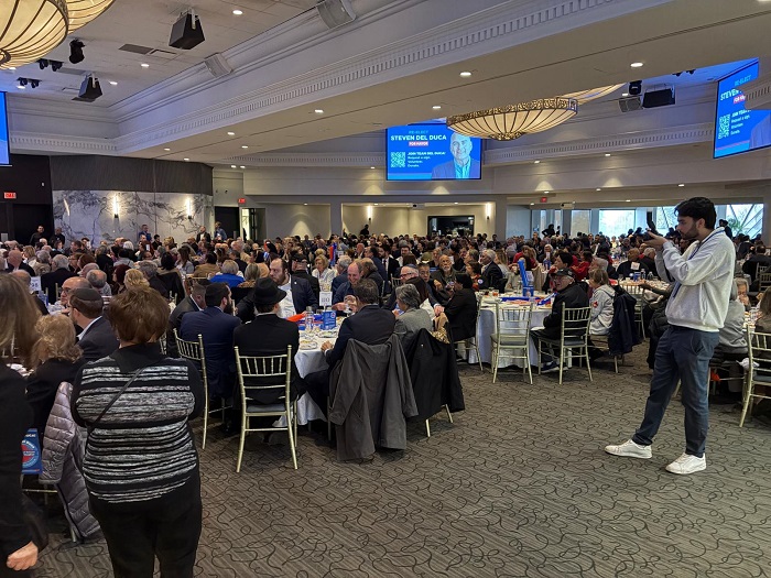 Large conference banquet hall crowded with round tables of attendees, a photographer taking pictures on the right side.