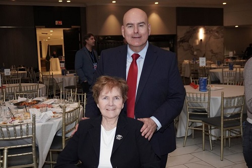 A bald man in a dark suit and red tie stands behind a seated woman at a banquet hall with round tables and chairs to celebrate an event.